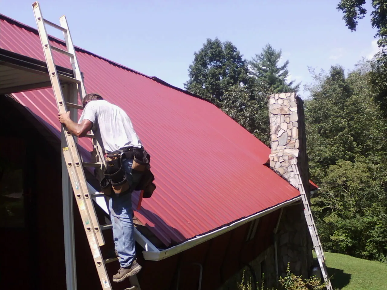 Roofer on a ladder beside a red metal roof and stone chimney — Soffit & Fascia Repair by Roofing Brothers in Colonial Beach
